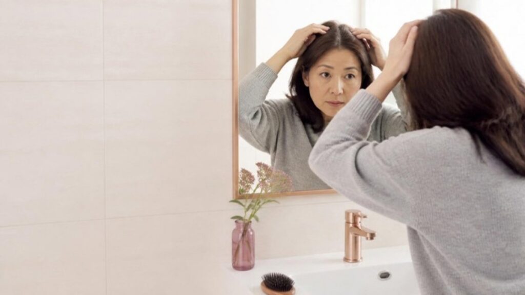 洗面台の鏡の前で髪の分け目を分けて、髪の薄さを確かめている50代の女性の画像