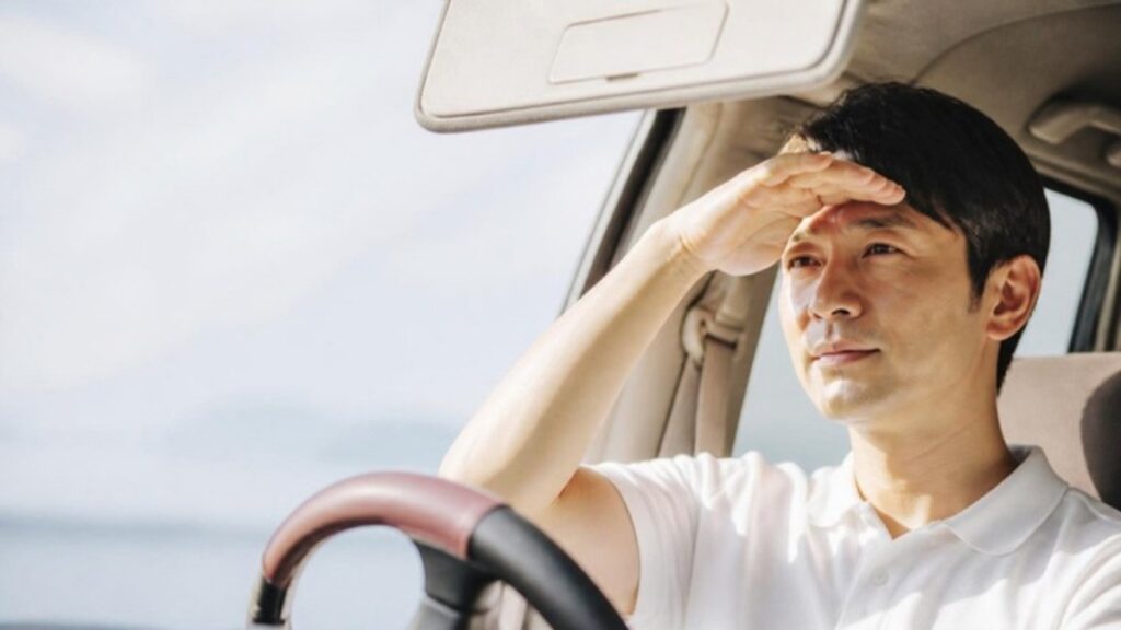 車の運転席で強い日差しに目を細め、手で顔に日よけをつくっている男性の画像